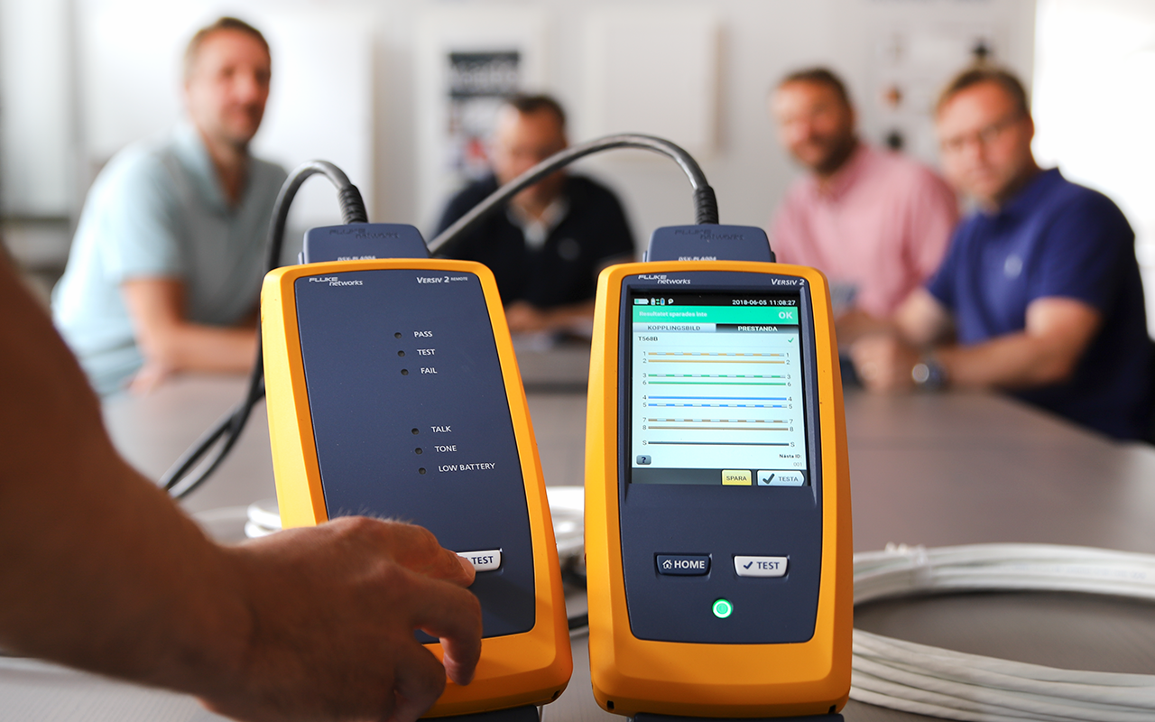 Zwei Fluke Messgeräte mit farbigem Display im Vordergrund. Im Hintergrund sind Mitarbeiter an einem Besprechungstisch versammelt.